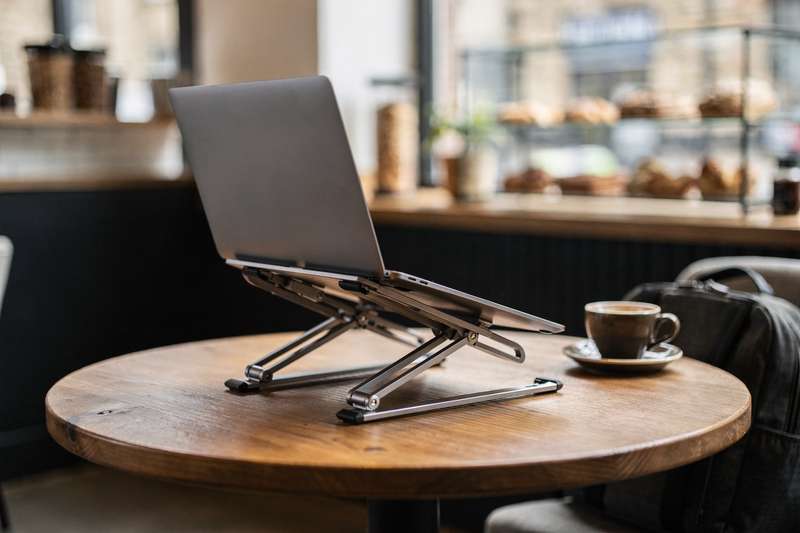 Uomo seduto al tavolo del bar lavora al laptop posizionato su un supporto pieghevole in alluminio.
