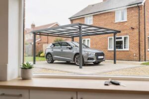 Una tettoia auto moderna in metallo grigio scuro protegge un'automobile parcheggiata in un vialetto di ghiaia inglese.